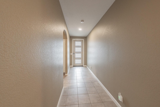 hall featuring arched walkways, light tile patterned flooring, a textured wall, and baseboards