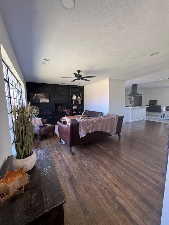 living room featuring visible vents, dark wood-type flooring, a brick fireplace, ceiling fan, and a textured ceiling