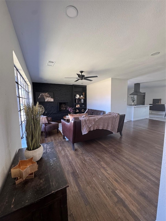 living area featuring a textured ceiling, visible vents, a ceiling fan, a brick fireplace, and dark wood finished floors