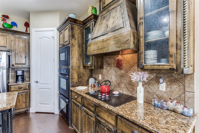 kitchen with glass insert cabinets, light stone countertops, black appliances, premium range hood, and backsplash