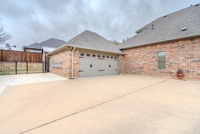 view of side of property with a garage, brick siding, fence, concrete driveway, and roof with shingles