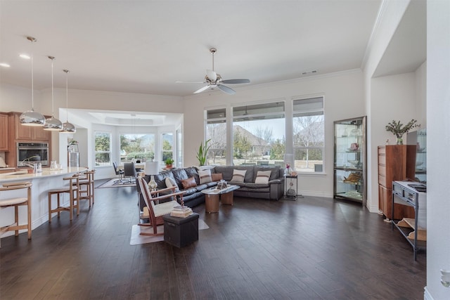 living room featuring dark wood-type flooring, visible vents, baseboards, a ceiling fan, and crown molding