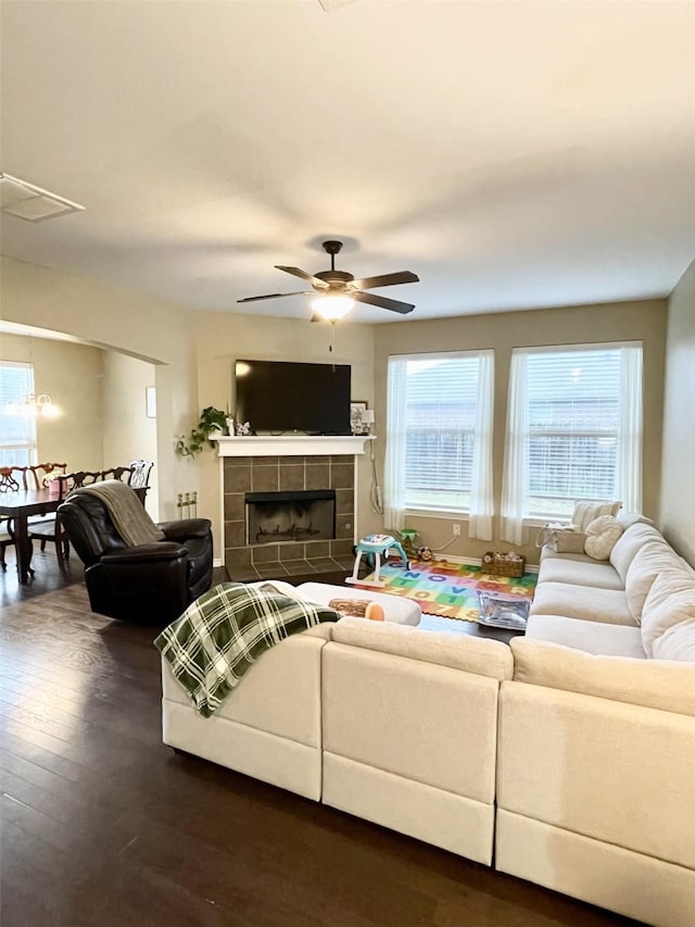 living room with ceiling fan, a tiled fireplace, visible vents, and dark wood-style flooring