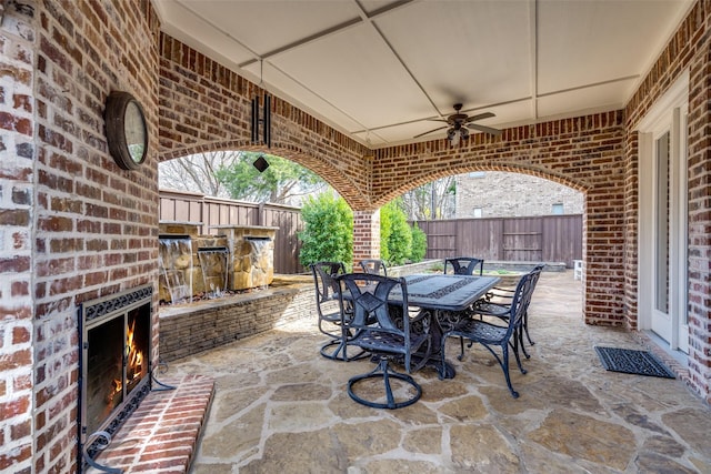 view of patio with outdoor dining space, a ceiling fan, fence, and an outdoor brick fireplace