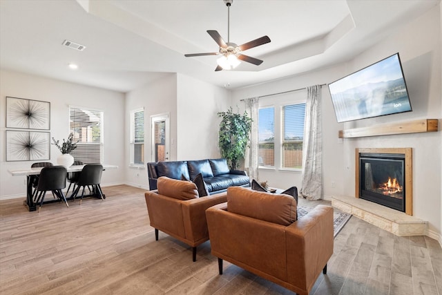 living area with visible vents, baseboards, light wood-type flooring, a glass covered fireplace, and a raised ceiling