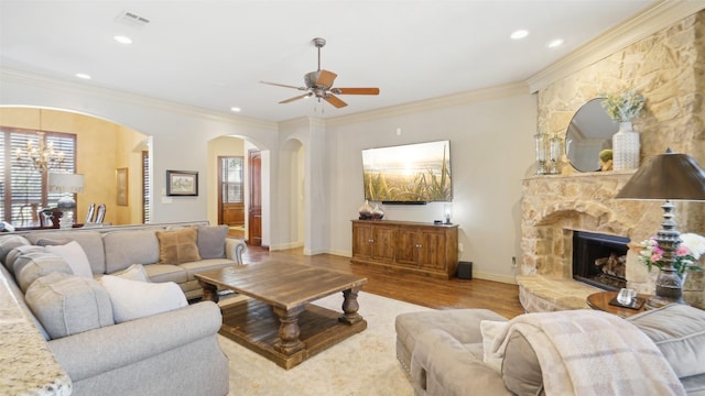 living room featuring wood finished floors, baseboards, a stone fireplace, crown molding, and ceiling fan with notable chandelier