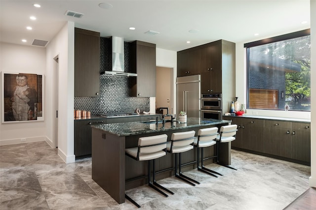 kitchen with visible vents, wall chimney range hood, an island with sink, stainless steel appliances, and a sink