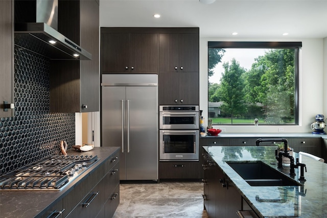 kitchen featuring a sink, dark stone counters, appliances with stainless steel finishes, wall chimney exhaust hood, and dark brown cabinets