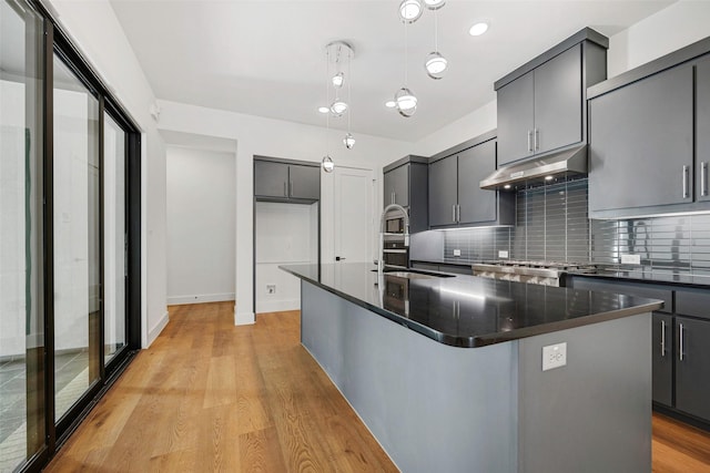 kitchen featuring under cabinet range hood, light wood-type flooring, tasteful backsplash, and decorative light fixtures