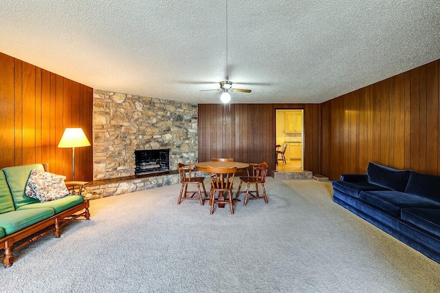 interior space with a textured ceiling, wooden walls, a stone fireplace, carpet flooring, and ceiling fan