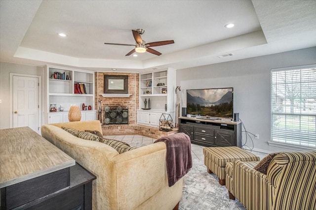 tiled living room with built in shelves, baseboards, ceiling fan, a raised ceiling, and a brick fireplace