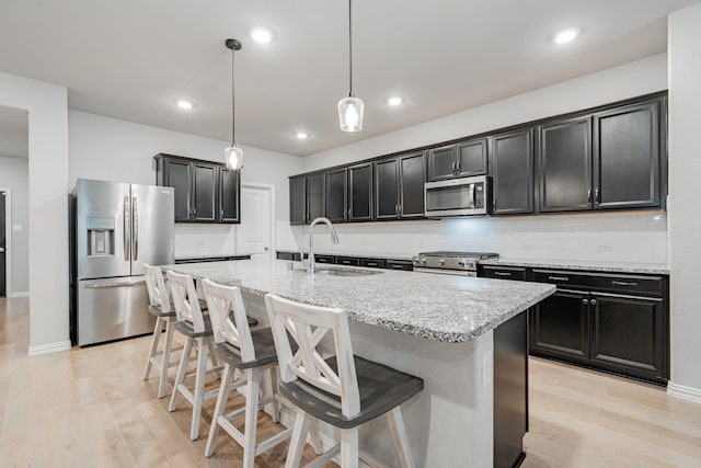 kitchen featuring an island with sink, light stone counters, appliances with stainless steel finishes, light wood finished floors, and decorative backsplash