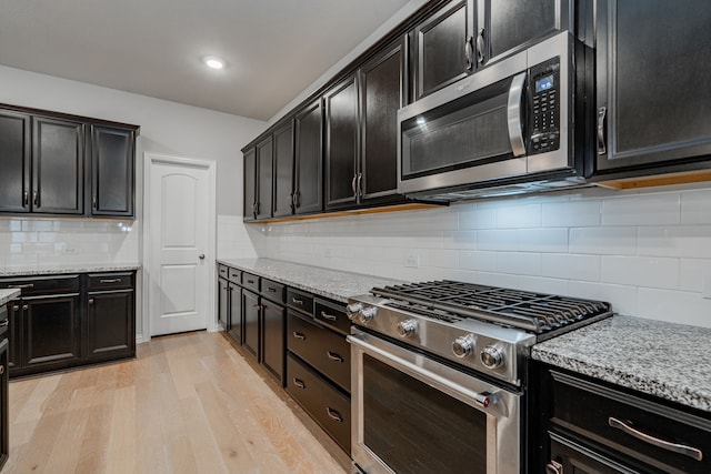 kitchen with dark cabinetry, light stone countertops, decorative backsplash, light wood-style floors, and appliances with stainless steel finishes