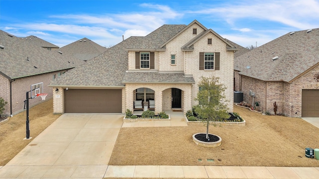 french country inspired facade featuring cooling unit, a shingled roof, concrete driveway, a garage, and brick siding