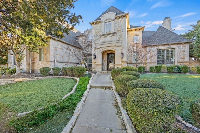 french country inspired facade with stone siding, a front yard, a chimney, and a shingled roof