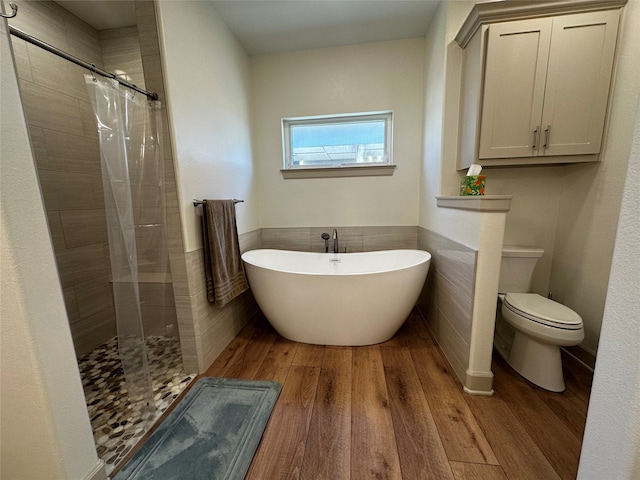 full bath featuring toilet, a freestanding bath, tiled shower, wood finished floors, and tile walls