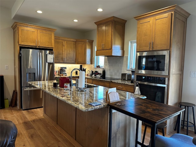 kitchen with brown cabinetry, stone counters, stainless steel appliances, light wood-style floors, and tasteful backsplash