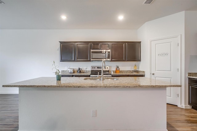 kitchen featuring light stone counters, wood finished floors, an island with sink, dark brown cabinets, and appliances with stainless steel finishes