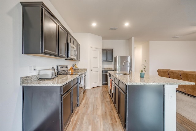 kitchen featuring light wood finished floors, open floor plan, a center island with sink, light stone counters, and appliances with stainless steel finishes