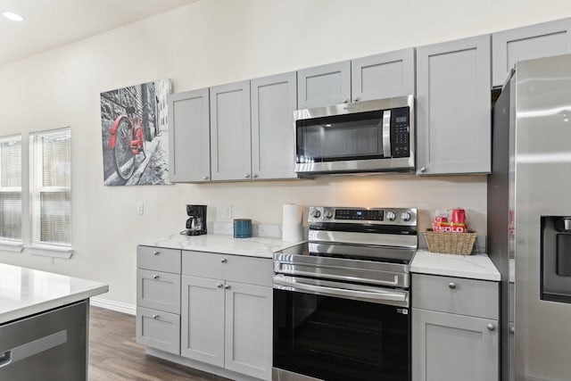 kitchen with gray cabinetry, light stone counters, wood finished floors, stainless steel appliances, and baseboards