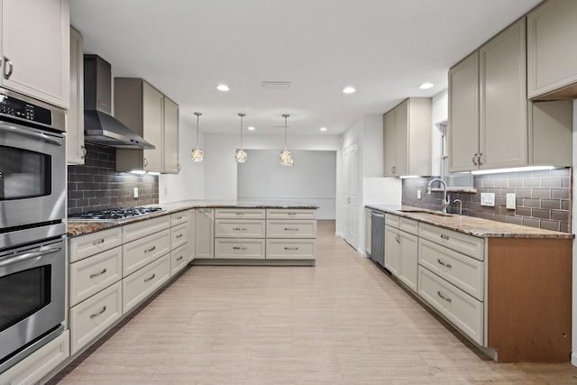 kitchen featuring light stone counters, a peninsula, a sink, appliances with stainless steel finishes, and wall chimney range hood