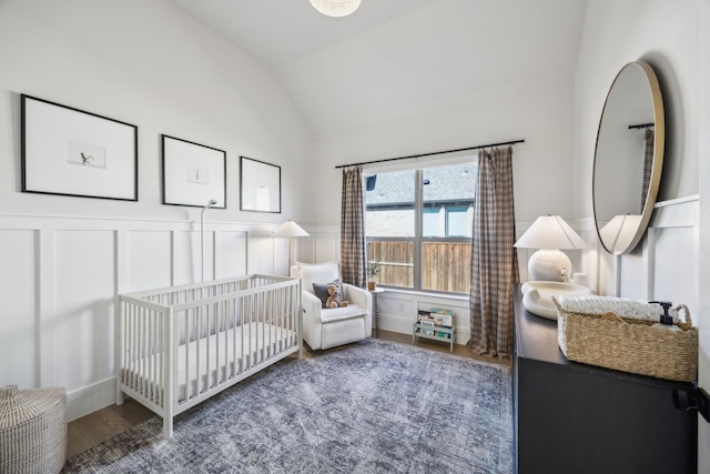 bedroom with wood finished floors, a wainscoted wall, vaulted ceiling, a nursery area, and a decorative wall