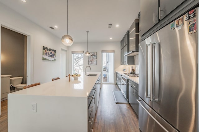 kitchen with a center island with sink, dark wood-style flooring, a sink, stainless steel appliances, and wall chimney exhaust hood