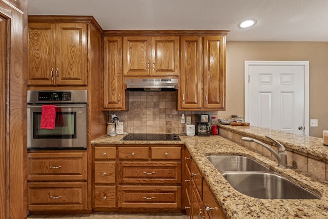 kitchen with oven, under cabinet range hood, brown cabinets, black electric cooktop, and a sink