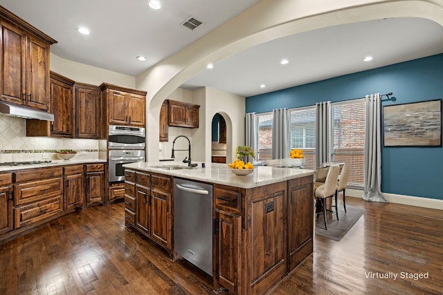 kitchen with a sink, under cabinet range hood, dark wood finished floors, stainless steel appliances, and decorative backsplash