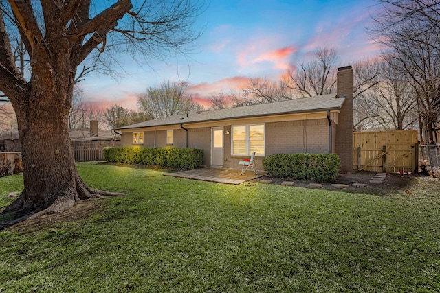 back of house at dusk featuring a gate, fence, a chimney, a lawn, and brick siding