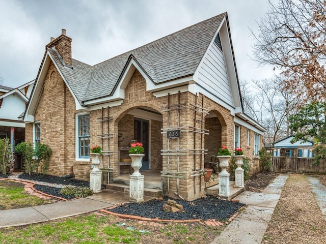 tudor home with a shingled roof, fence, brick siding, and a chimney