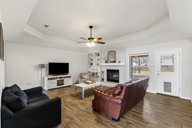 living room with wood finished floors, visible vents, baseboards, a tray ceiling, and a brick fireplace