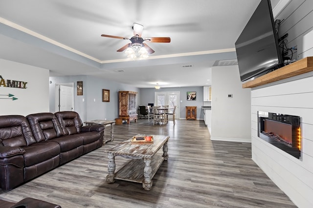 living area featuring ceiling fan, baseboards, a tray ceiling, wood finished floors, and a glass covered fireplace