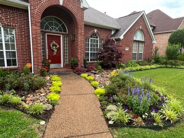 doorway to property featuring a yard, brick siding, and roof with shingles