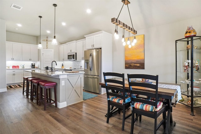 kitchen featuring dark wood-type flooring, a center island with sink, decorative backsplash, appliances with stainless steel finishes, and white cabinetry