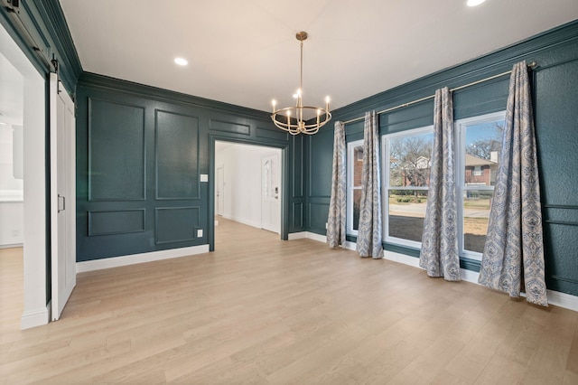 unfurnished dining area with light wood-style flooring, a barn door, crown molding, a decorative wall, and a chandelier