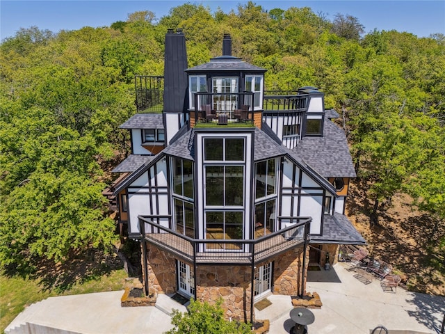 rear view of property featuring a shingled roof, a wooded view, stone siding, french doors, and driveway