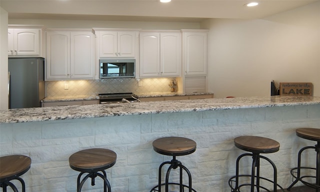 kitchen with backsplash, light stone counters, appliances with stainless steel finishes, a kitchen breakfast bar, and white cabinets