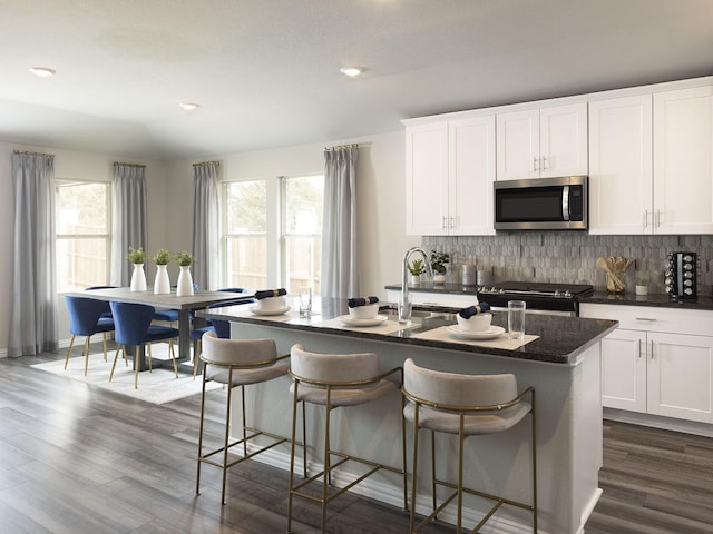 kitchen featuring stainless steel microwave, backsplash, range with gas cooktop, a center island with sink, and dark wood-style floors