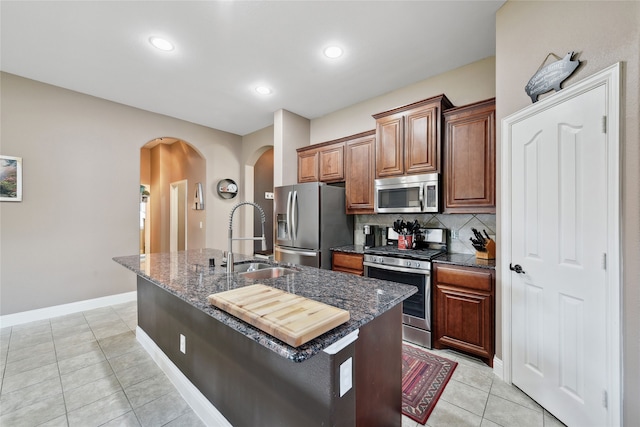521 Eastbrook Dr, Anna, TX 75409 - kitchen with stainless steel appliances, arched walkways, light tile patterned floors, a kitchen island with sink, and dark stone counters