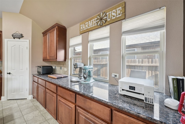 kitchen with dark stone countertops, light tile patterned floors, decorative backsplash, vaulted ceiling, and brown cabinets