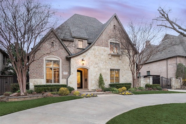 french country style house with stone siding, brick siding, a shingled roof, and fence