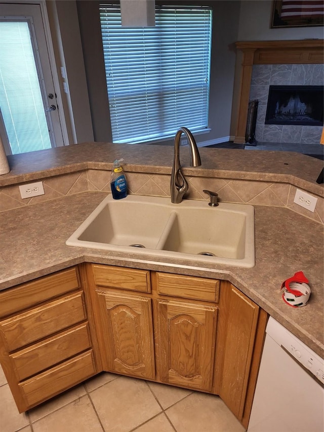 kitchen with light tile patterned floors, white dishwasher, a high end fireplace, a sink, and brown cabinets