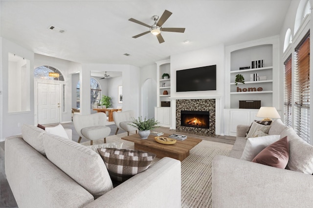 living area featuring visible vents, ceiling fan, a tiled fireplace, built in features, and wood finished floors