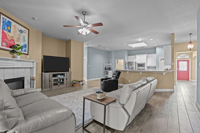 living room featuring a ceiling fan, baseboards, a skylight, a tiled fireplace, and light wood-type flooring