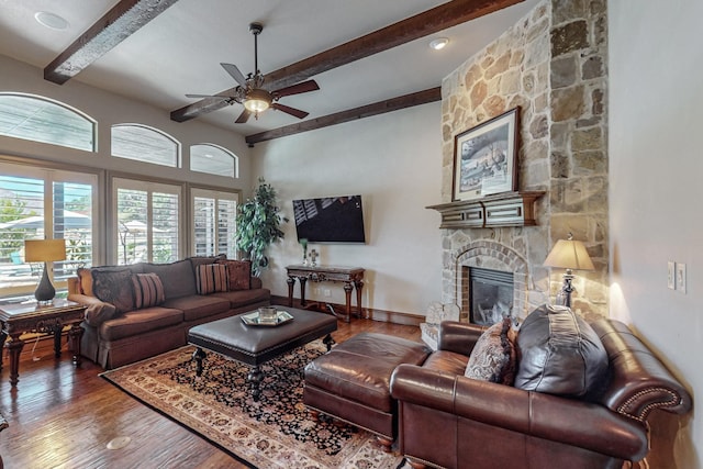 living room featuring ceiling fan, baseboards, beamed ceiling, a stone fireplace, and wood finished floors