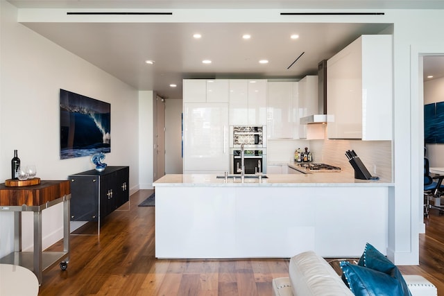 kitchen featuring stainless steel gas stovetop, wall chimney range hood, a sink, a peninsula, and white cabinetry