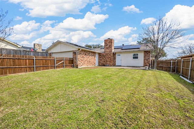 rear view of property with roof mounted solar panels, a fenced backyard, a yard, brick siding, and a chimney