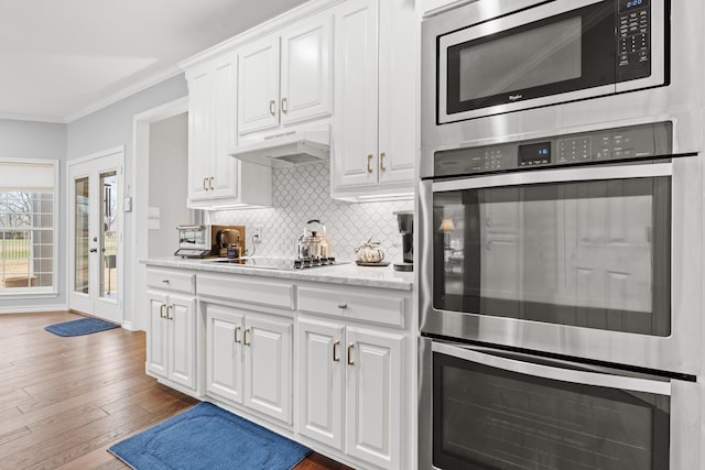 kitchen with crown molding, under cabinet range hood, decorative backsplash, appliances with stainless steel finishes, and wood-type flooring