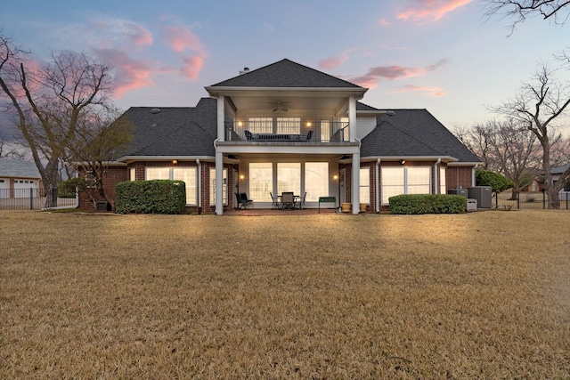 back of property at dusk featuring fence, a yard, a balcony, ceiling fan, and brick siding